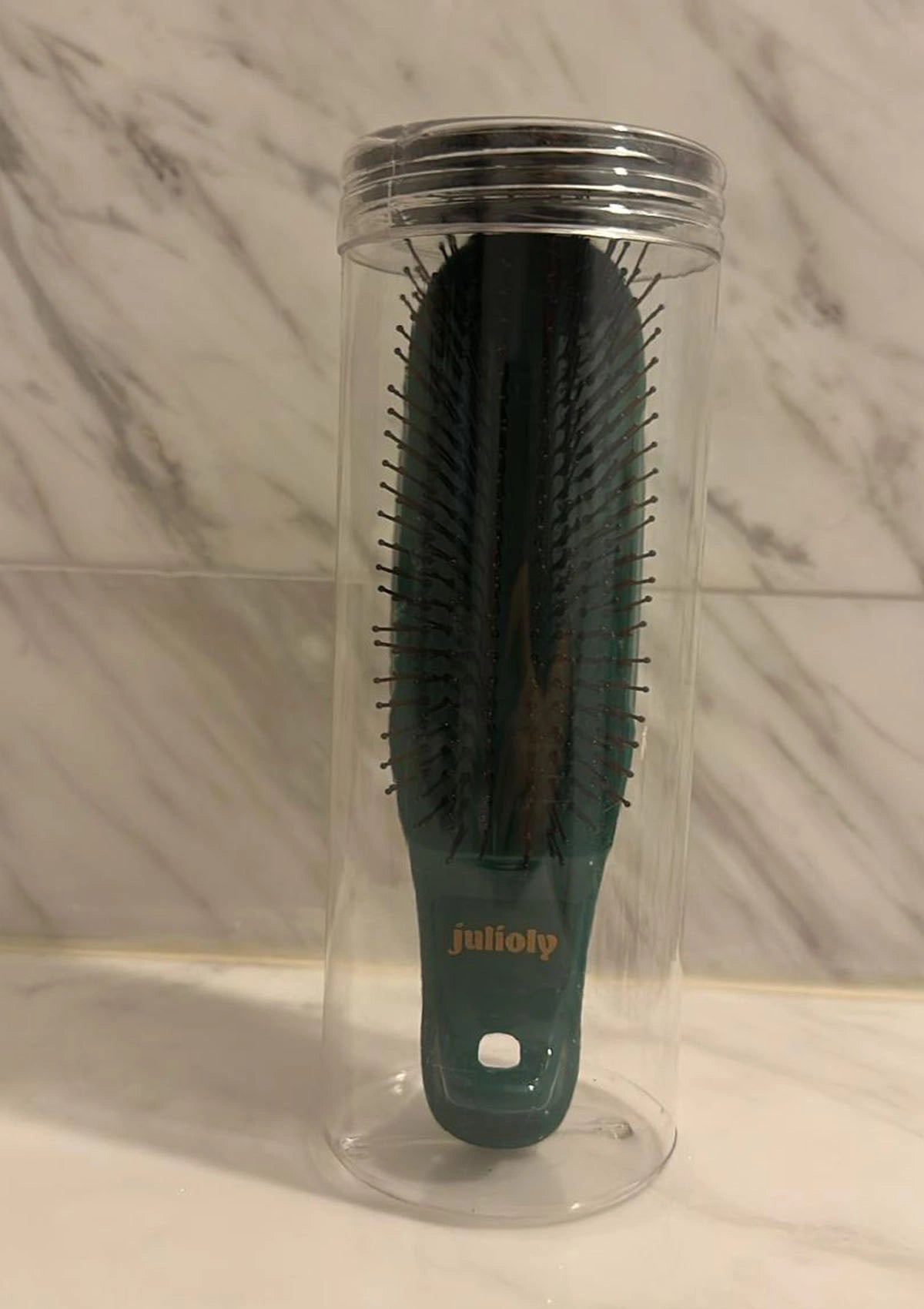 Black hairbrush with green handle in a clear plastic stand against a marble wall.